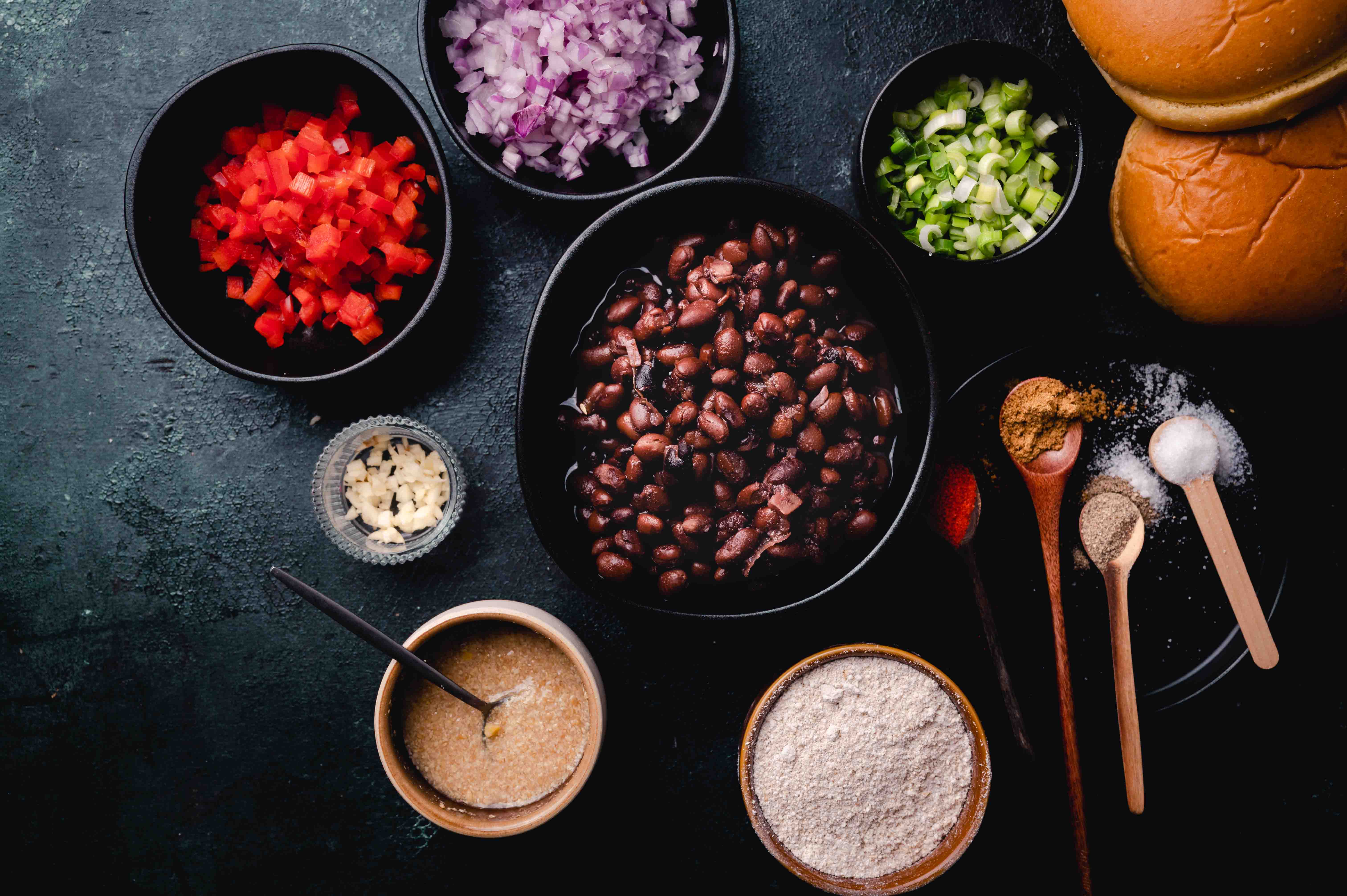 Overhead view of a kitchen counter with ingredients for cooking, featuring bowls of beans, chopped vegetables, spices, and bread on a dark surface.