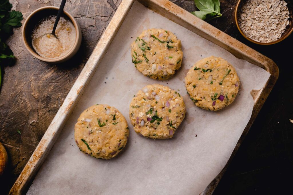Four homemade veggie burgers on a parchment-lined baking sheet, with oatmeal and herbs visible, accompanied by a bowl of oat flakes and a spoon.