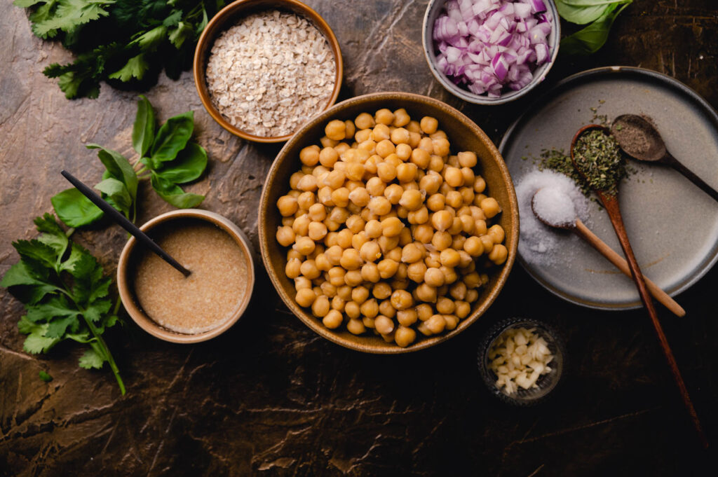 Overhead view of a bowl of chickpeas surrounded by ingredients including oats, chopped onions, garlic, and spices on a dark textured surface.