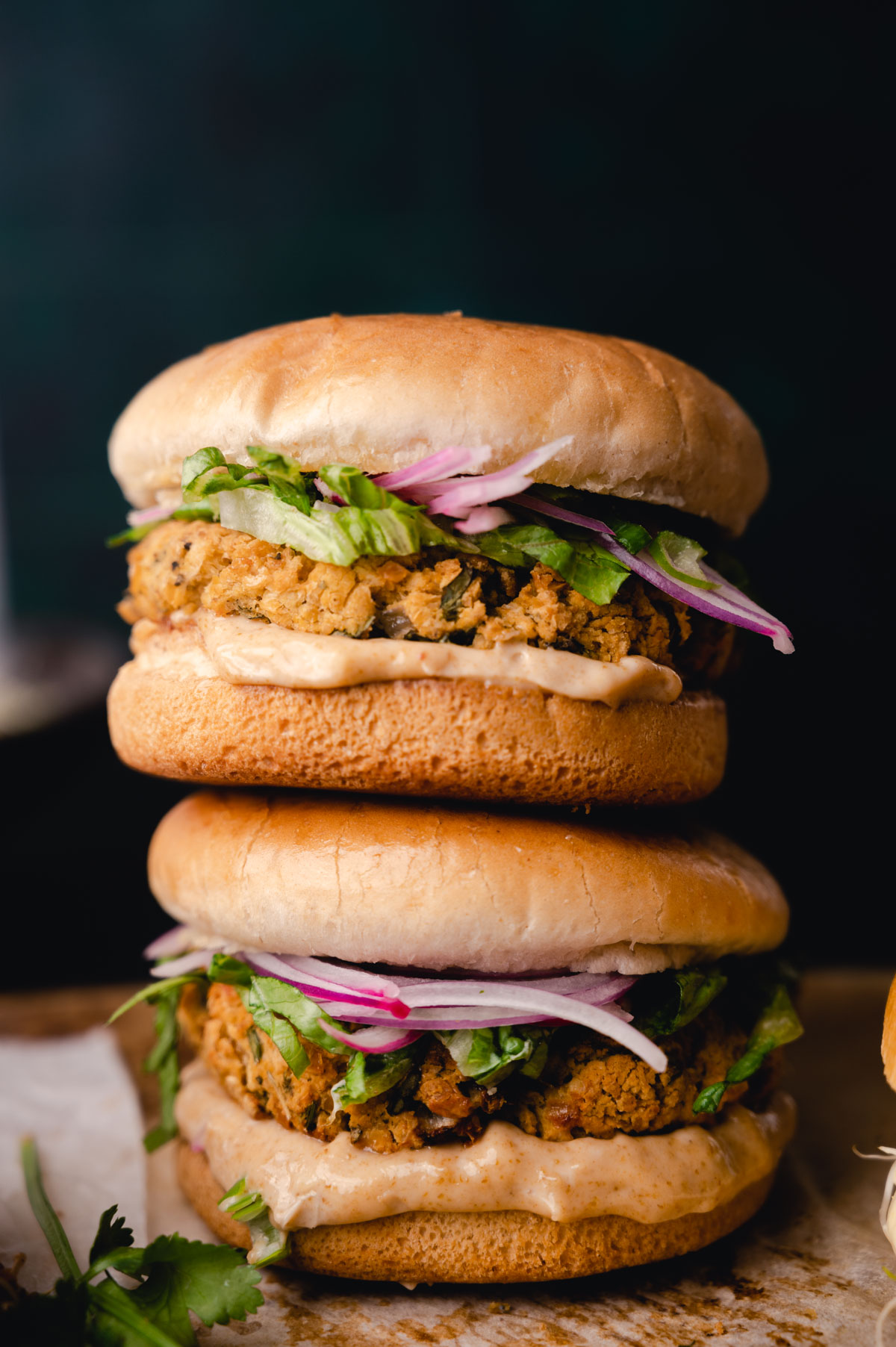 Two stacked fried chicken sandwiches with lettuce, onions, and sauce on sesame buns, on a dark background.