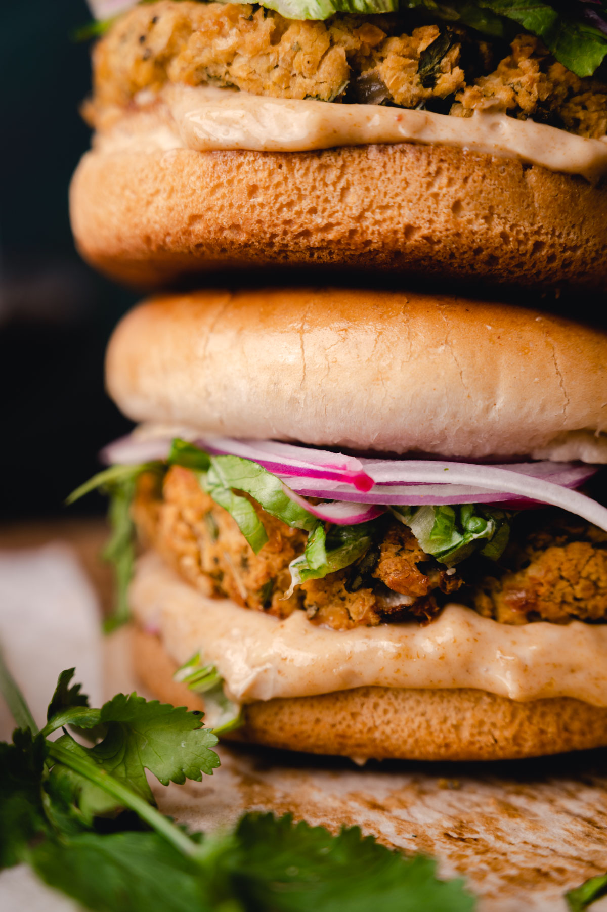 Close-up of two stacked falafel burgers with creamy sauce, fresh cilantro, and sliced red onions in sesame seed buns.