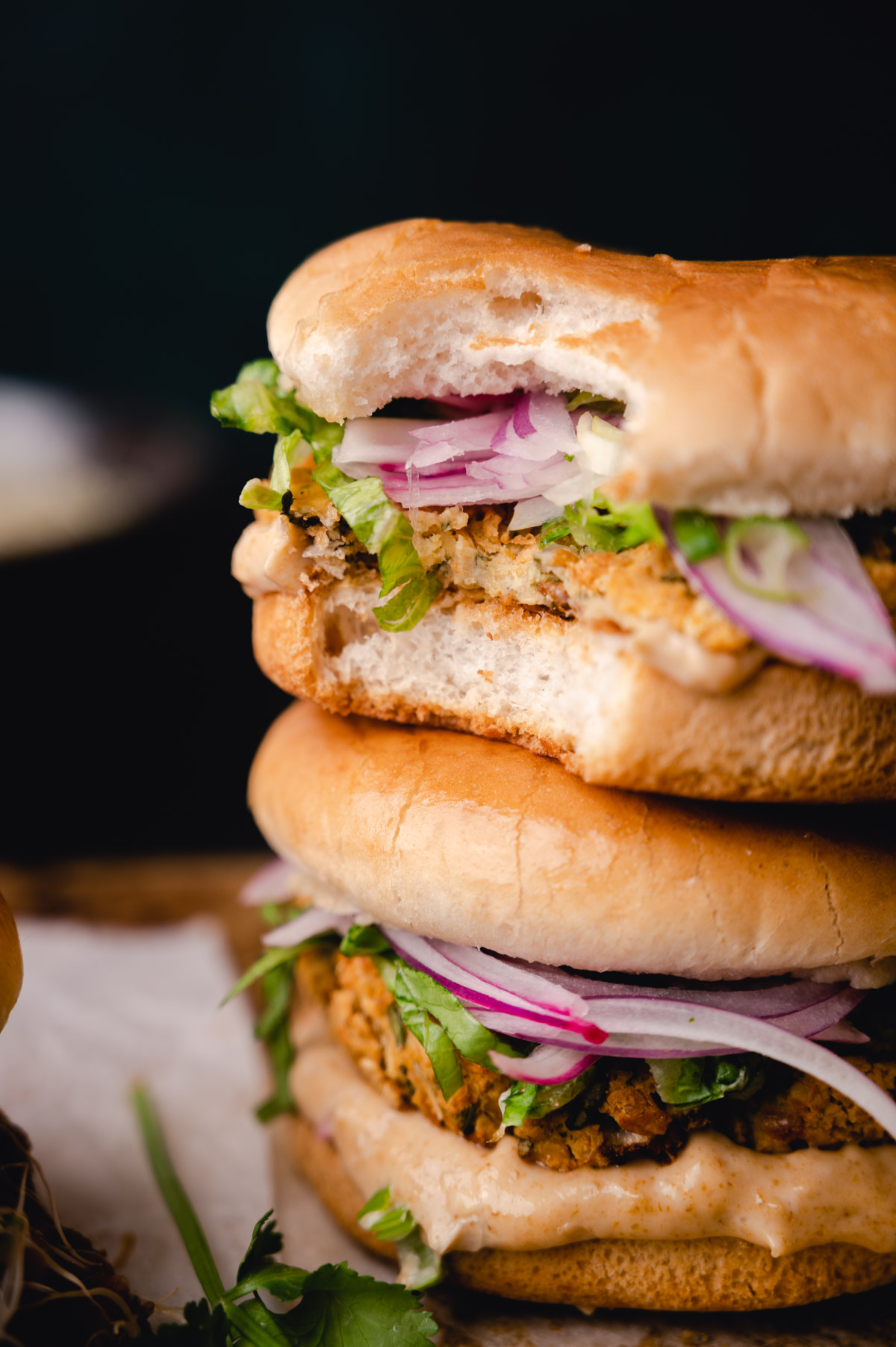 Close-up of a double-layer veggie burger with red onions, lettuce, and sauce on a bun, set against a dark background.