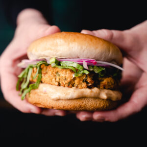Two hands holding a chickpea burger with lettuce, red onions, and sauce in a bun.