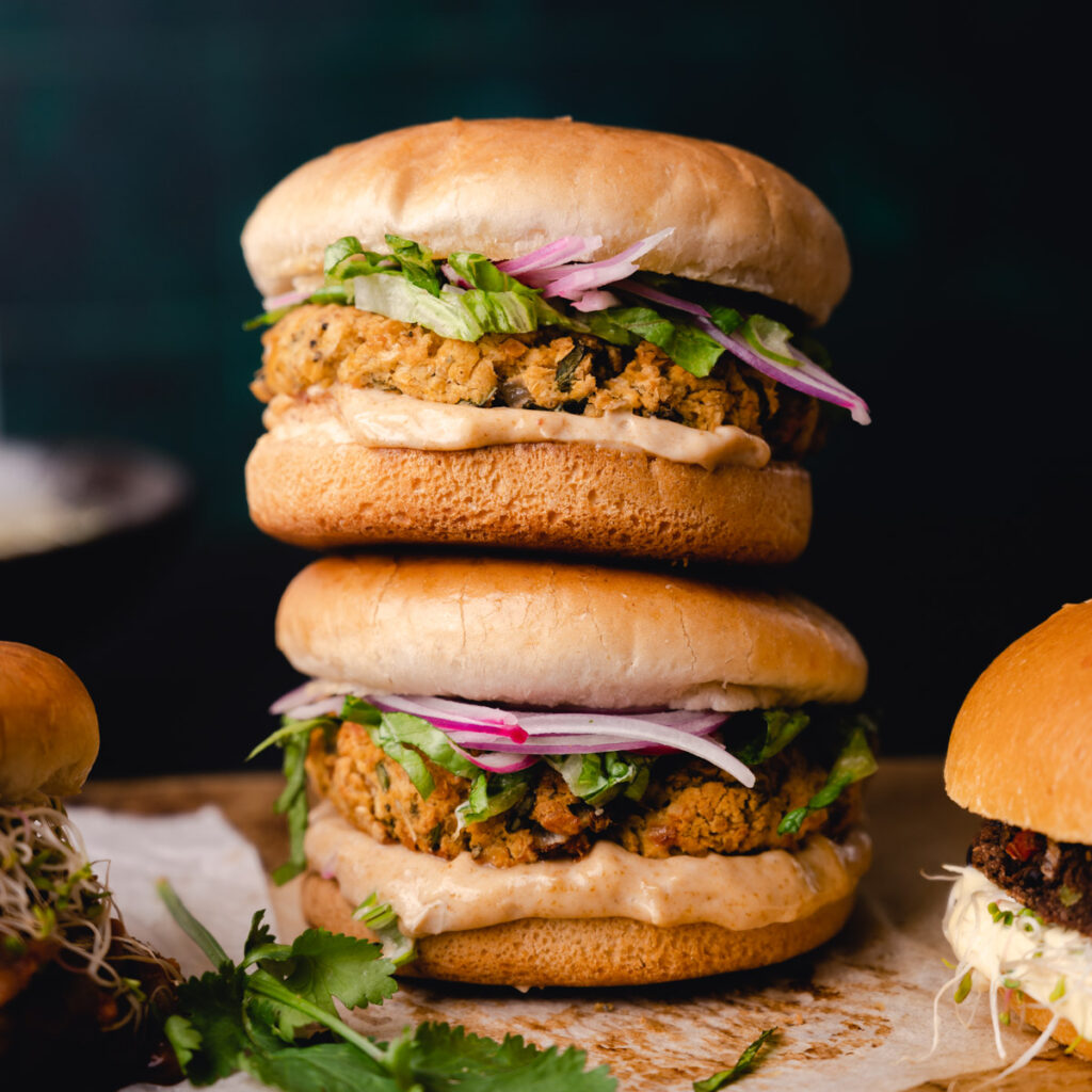 Two stacked falafel burgers with red onion and cilantro on sesame buns, presented on a wooden surface with moody lighting.