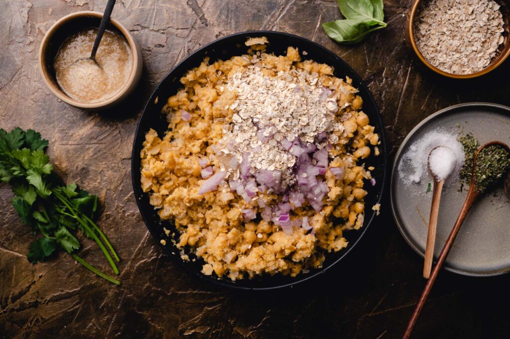 A bowl of lentil risotto topped with onions and oats, surrounded by ingredients like herbs, spices, and a sauce on a dark, textured surface.