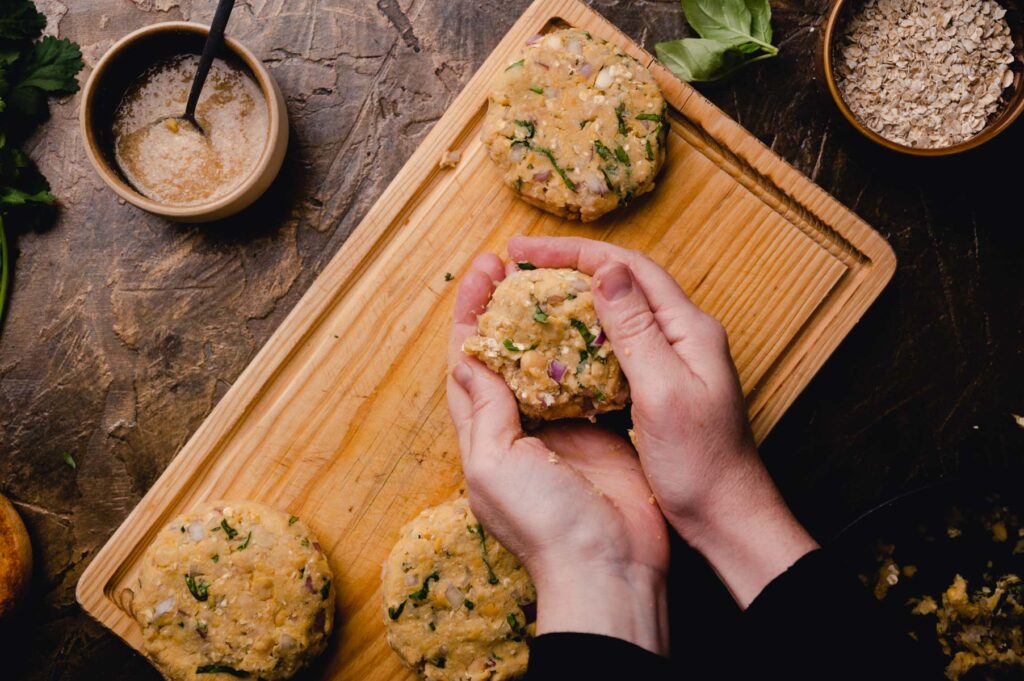 Person shaping chickpea patties on a wooden board with ingredients and utensils around on a dark table.