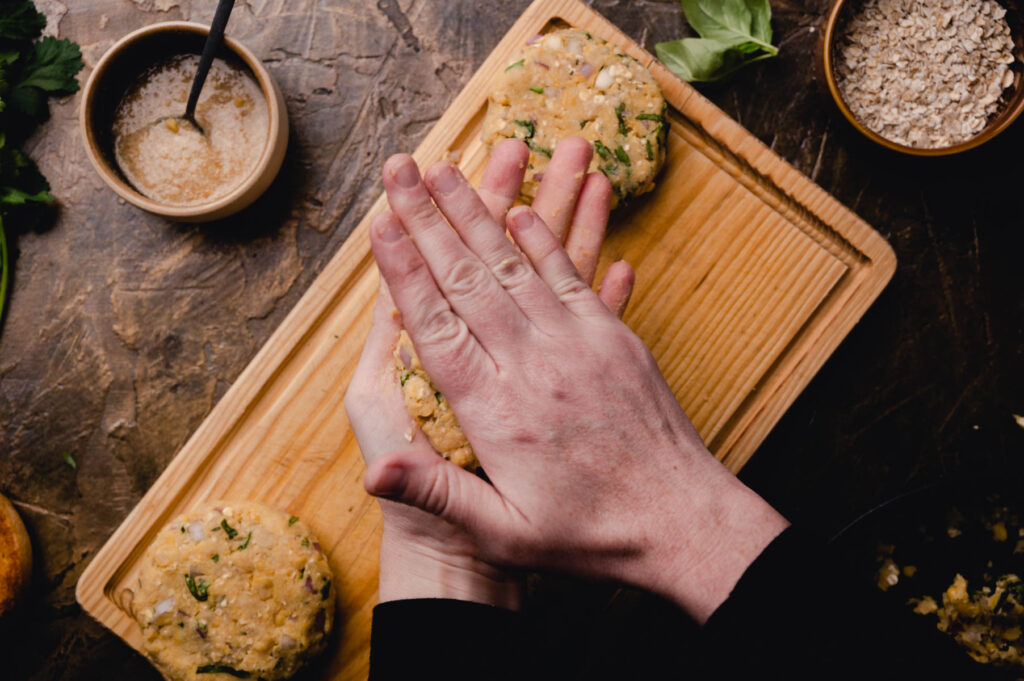 A person pressing down on chickpea mixture on a wooden cutting board, surrounded by ingredients like oats and herbs.