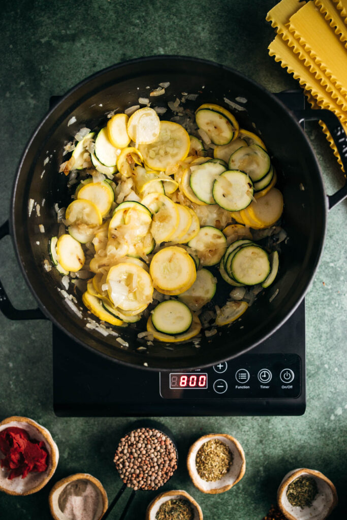 Sliced zucchini and onions cooking in a black electric pressure cooker with a digital display reading 8:00, surrounded by spices and pasta.