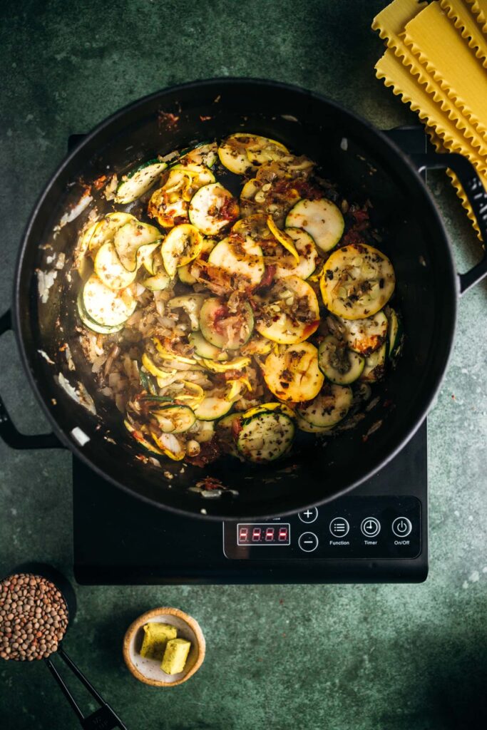 Top view of a skillet meal with sliced zucchini and other vegetables cooking on an induction cooker, with ingredients like pasta and spices nearby.