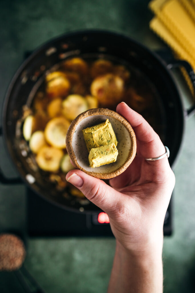 A person holding a small bowl of cubed herb butter over a pot of cooking sliced potatoes.