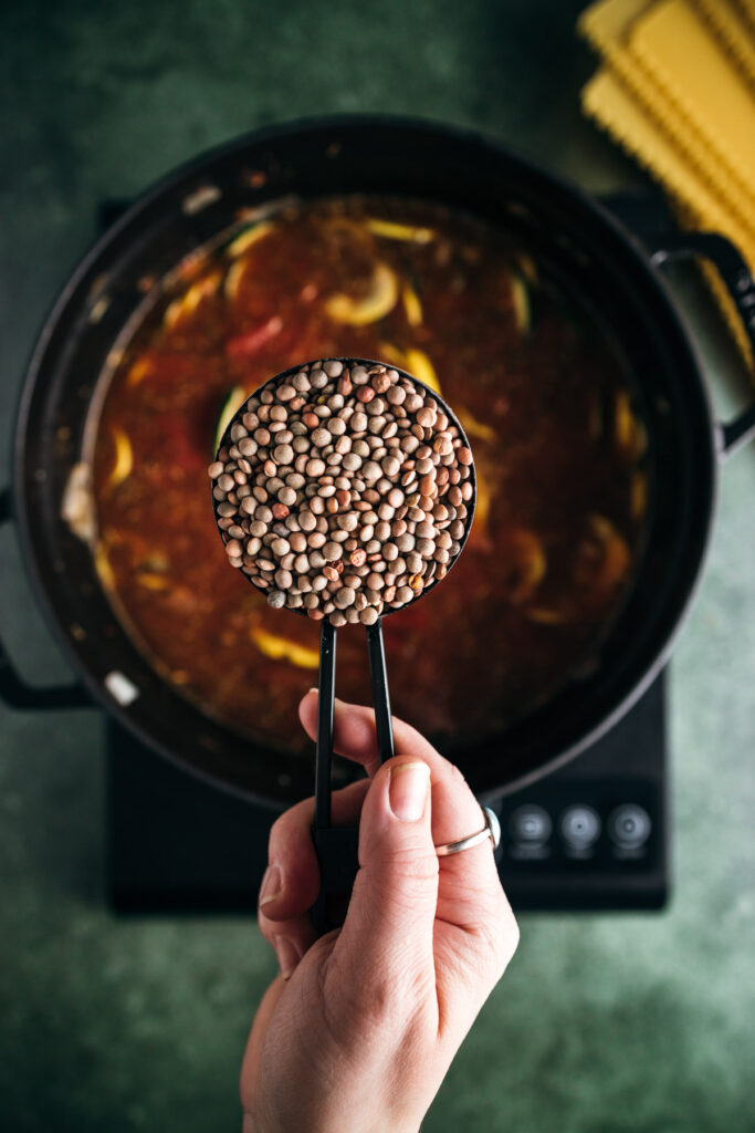 A person's hand holding a ladle filled with lentils over a pot of soup on an induction stove.