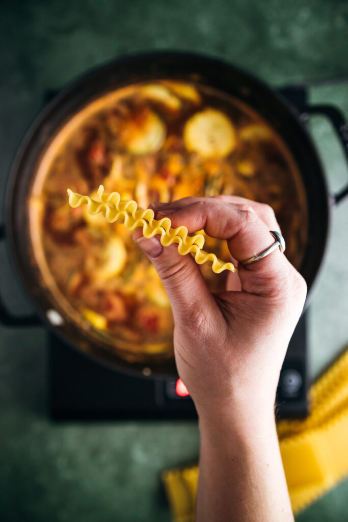 A hand holding a single piece of fusilli pasta over a pot of simmering stew with vegetables, shot from an aerial view.