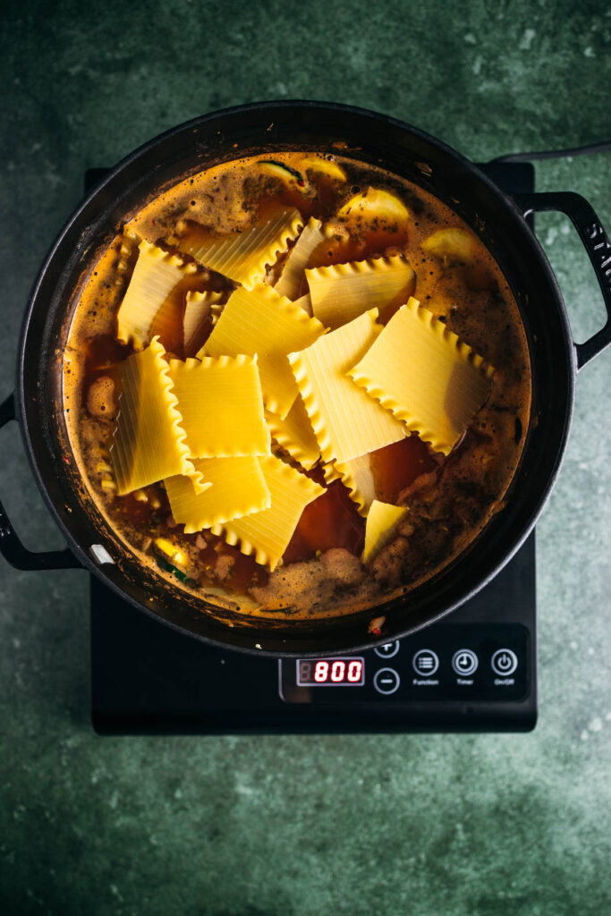 Top-down view of large pasta sheets cooking in a broth inside an electric pot set to 800 degrees displayed on its digital panel.