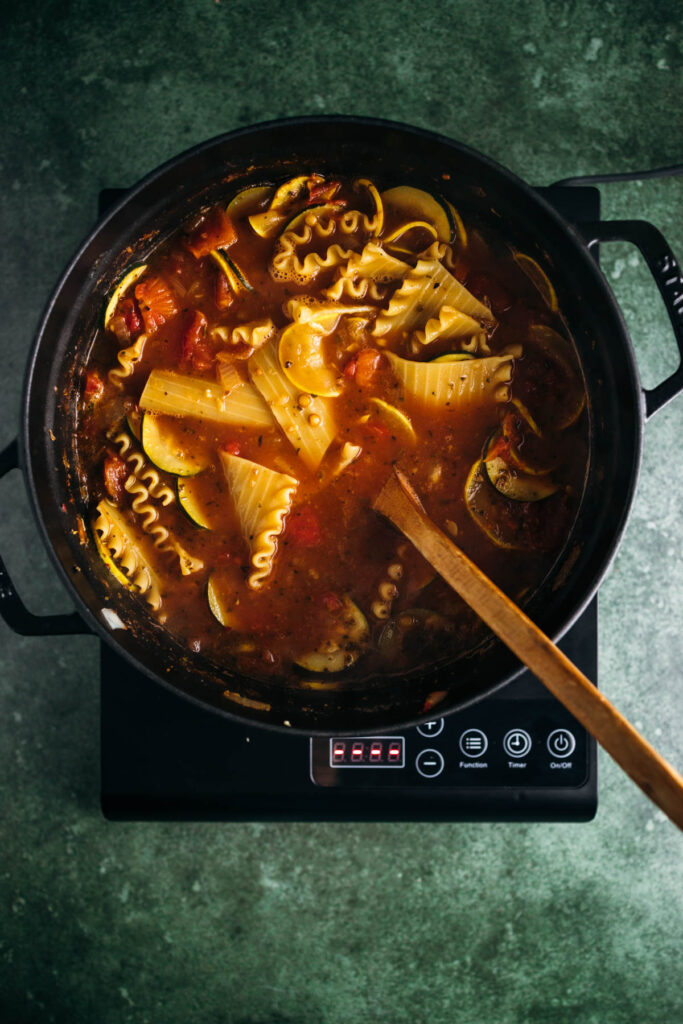 A pot of tomato-based pasta soup with mixed vegetables and spiral pasta, cooking on an induction cooker.