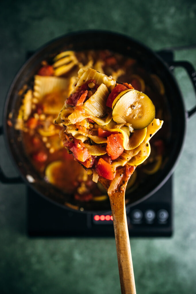 Wooden spoon lifting a serving of stew with vegetables and pasta from a pot on an induction cooker.