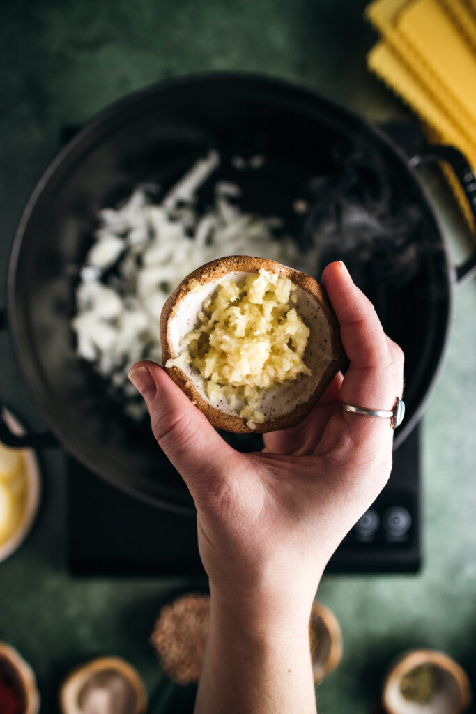 A person's hand holding a half of a baked potato filled with garlic mashed potatoes over a skillet with diced onions on a green surface.