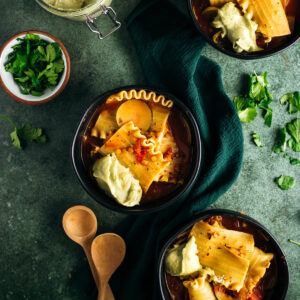 Overhead view of two bowls of lasagna soup garnished with ricotta cheese, served with fresh herbs on a textured green surface.