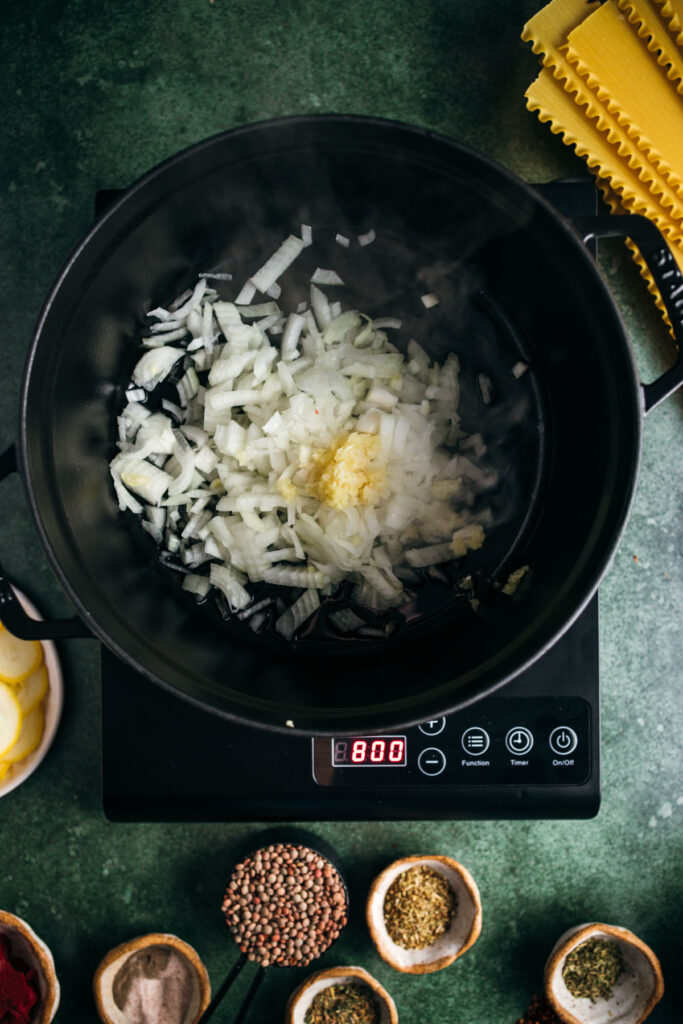 Chopped onions and garlic sautéing in a black induction cooker, with various spices and pasta arranged around it.