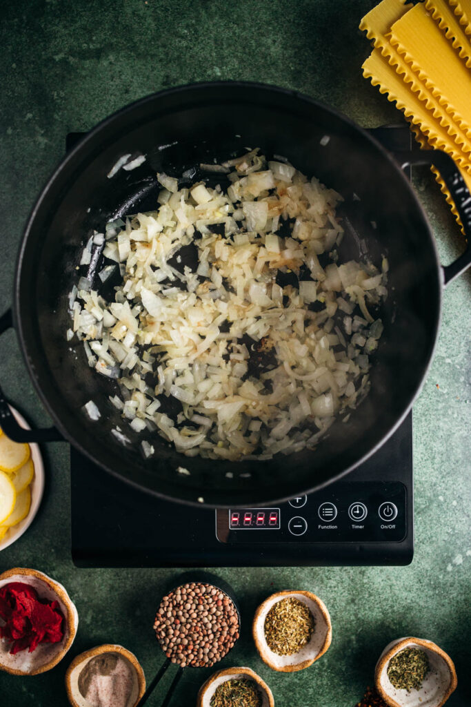 Chopped onions sautéing in a black skillet on a digital induction cooktop, surrounded by various spices and uncooked pasta on a green countertop.