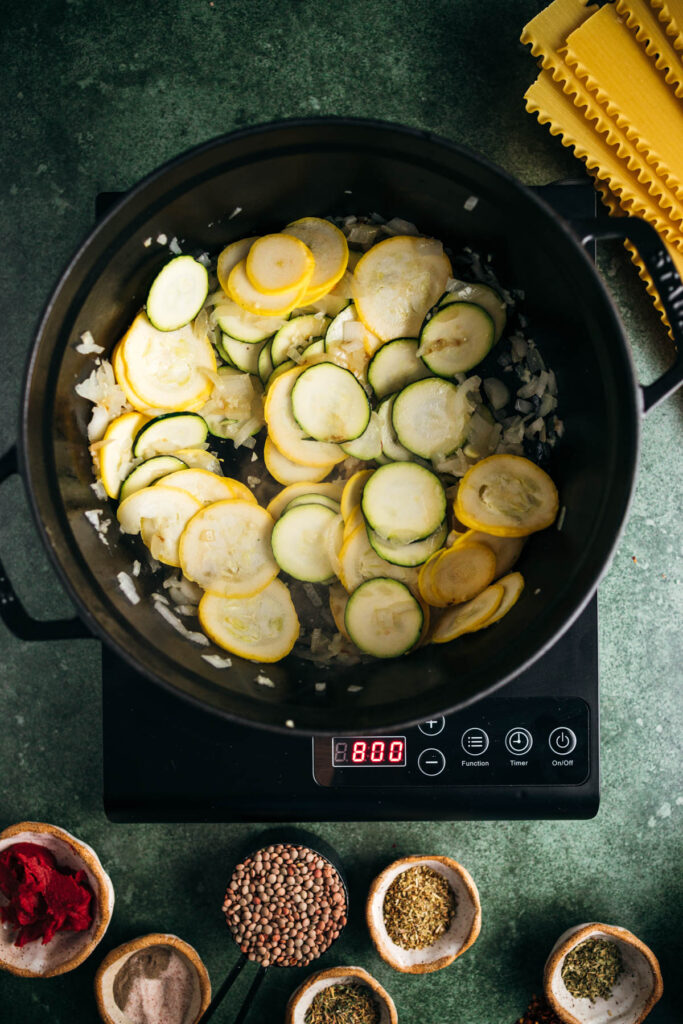 Top view of a cooking pot on an induction cooker with sliced zucchini and onions, surrounded by various spices and uncooked pasta.