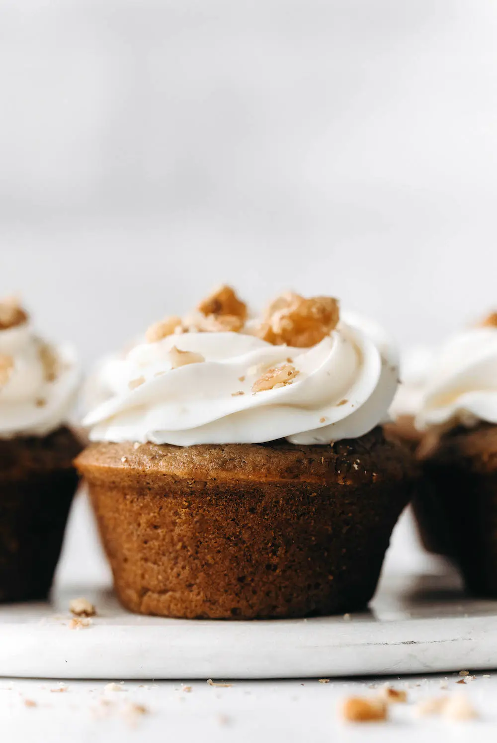 Three vegan carrot cake muffins with cream cheese frosting and crushed nuts on top, displayed on a white surface.