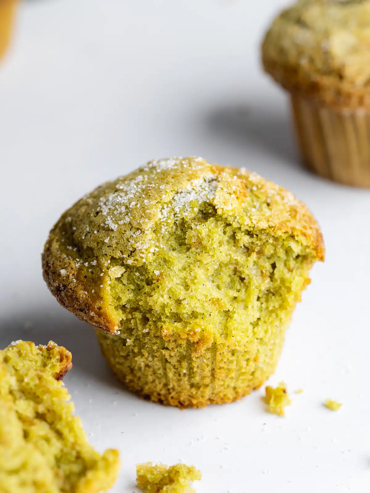 Close-up of a vegan green tea muffin with a dusting of powdered sugar, with another muffin and crumbs in the background.