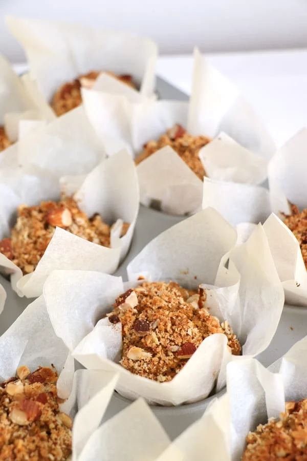 Freshly baked vegan muffins in white parchment paper liners, sprinkled with nuts and grains, displayed in a closeup setting.