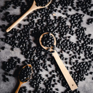 Three wooden spoons filled with black beans rest on a surface scattered with more black beans.