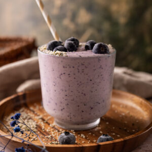 A glass of blueberry smoothie topped with whole blueberries and hemp seeds, served on a wooden tray with scattered blue flowers.