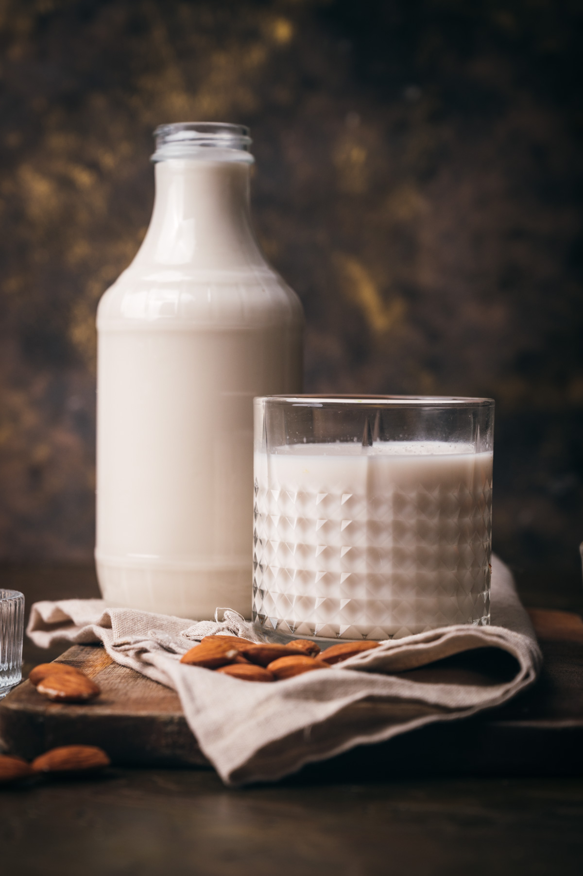 A bottle and a glass filled with milk on a wooden table, accompanied by almonds and a cloth, against a dark backdrop.
