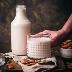 A person holding a glass of almond milk beside a bottle and a bowl of almonds on a cloth-covered table.
