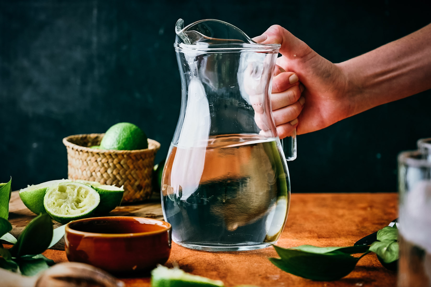 A person's hand holding a clear glass pitcher of water on a wooden table, surrounded by halved limes and a bowl of salt.