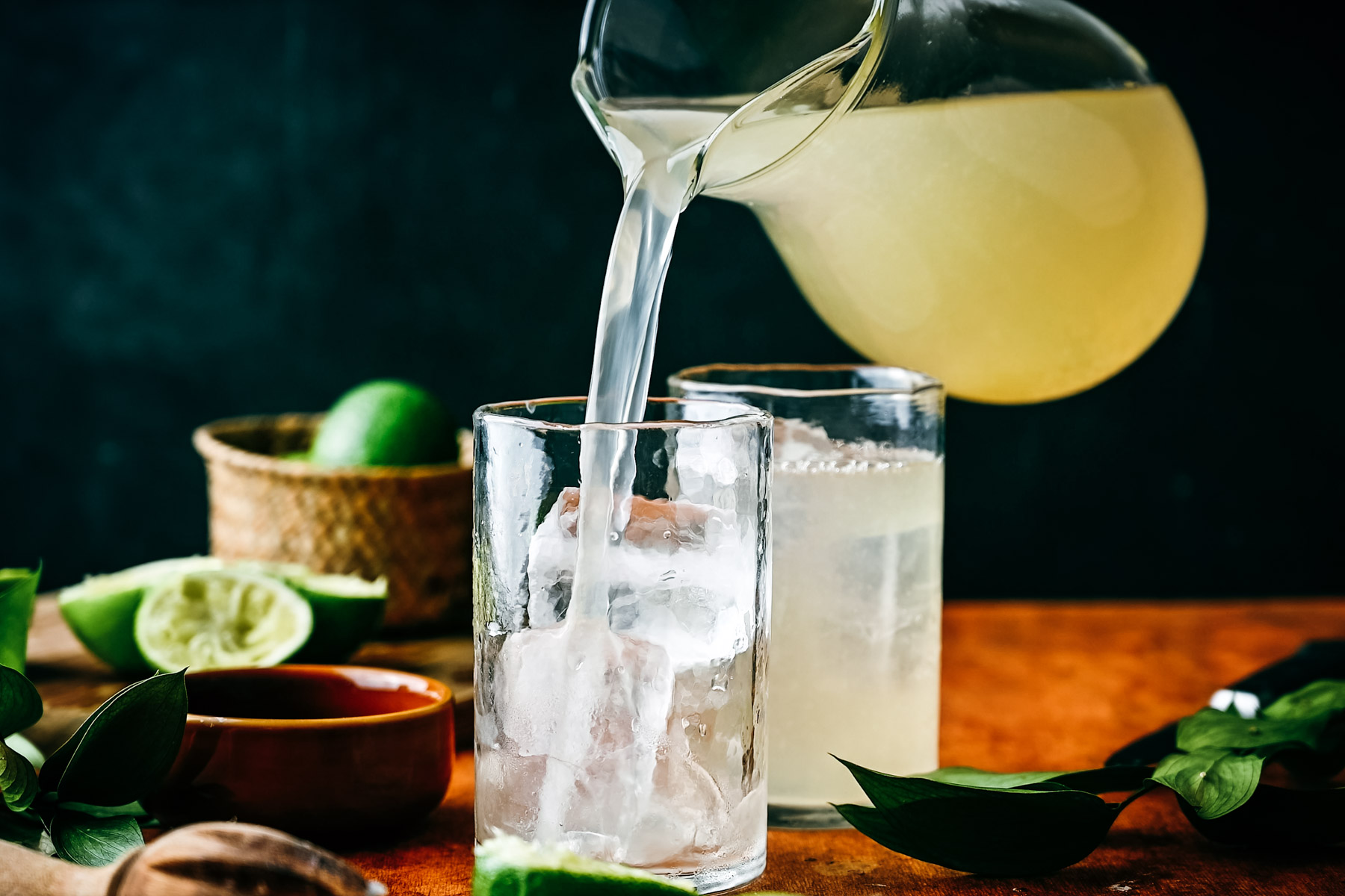 Pouring a clear, citrus drink into an ice-filled glass on a rustic table, surrounded by limes and fresh herbs.
