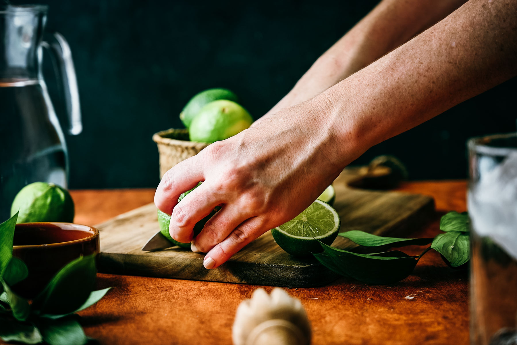 A person slicing a lime on a wooden cutting board, surrounded by fresh greens and a pitcher of water in the background.