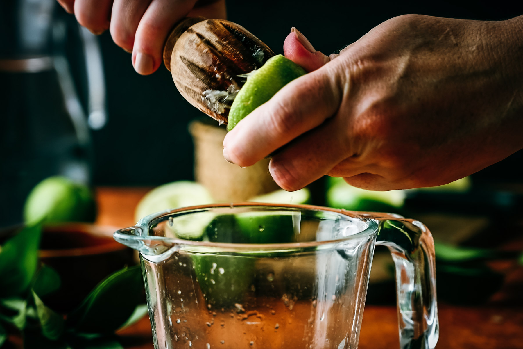 Hands squeezing lime juice into a glass pitcher using a wooden citrus reamer, with fresh limes scattered around.