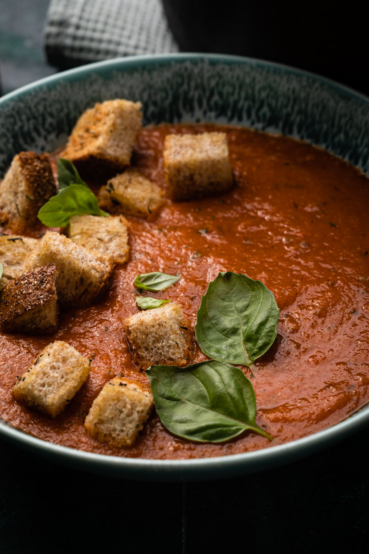 A bowl of tomato soup garnished with basil leaves and topped with croutons, served on a dark surface.