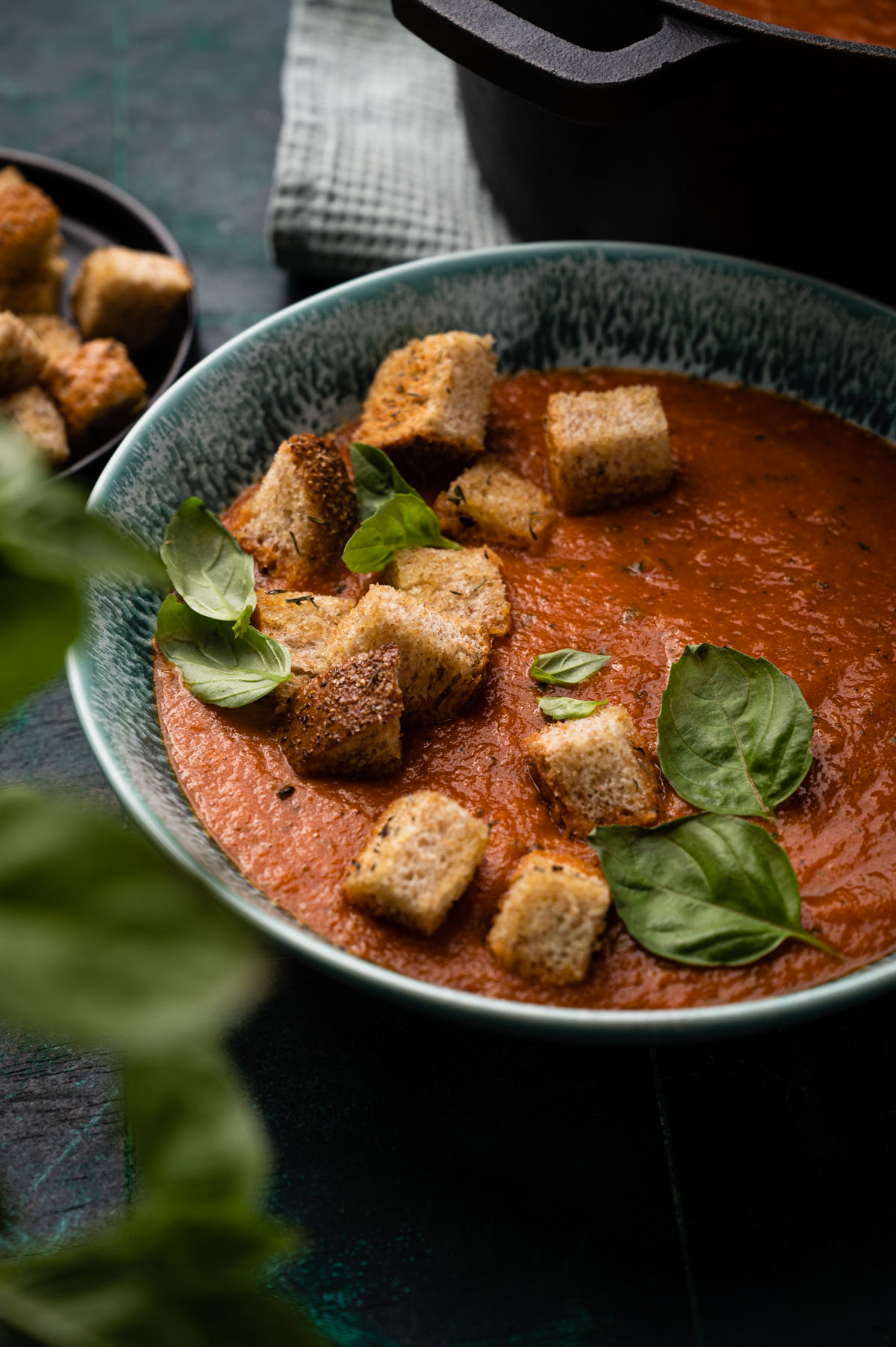 A bowl of tomato soup garnished with fresh basil and croutons, next to a cast iron pot, on a dark, textured surface.