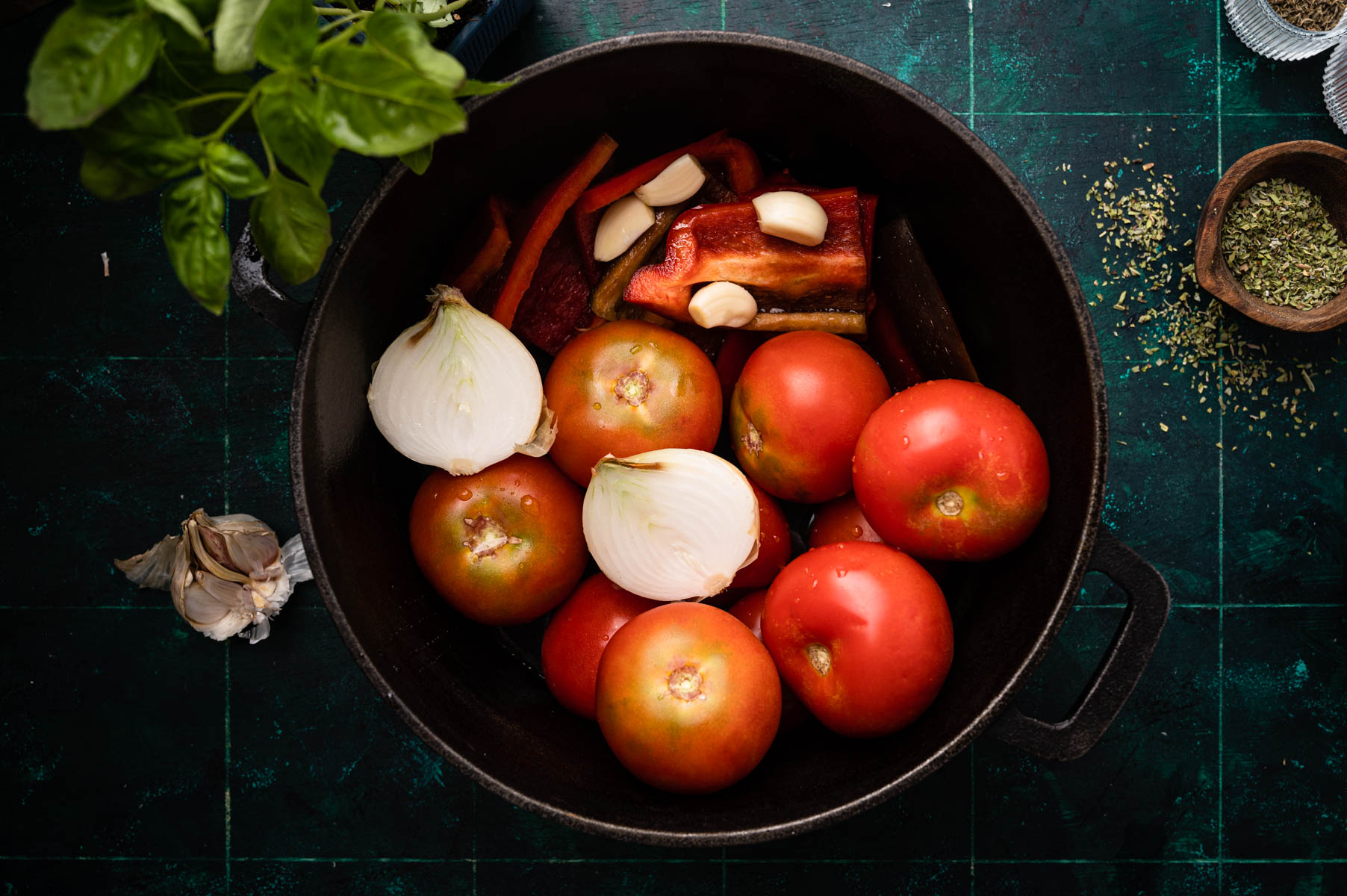 A cast iron skillet containing fresh tomatoes, onions, garlic, and red peppers, ready for cooking, on a dark, textured surface.
