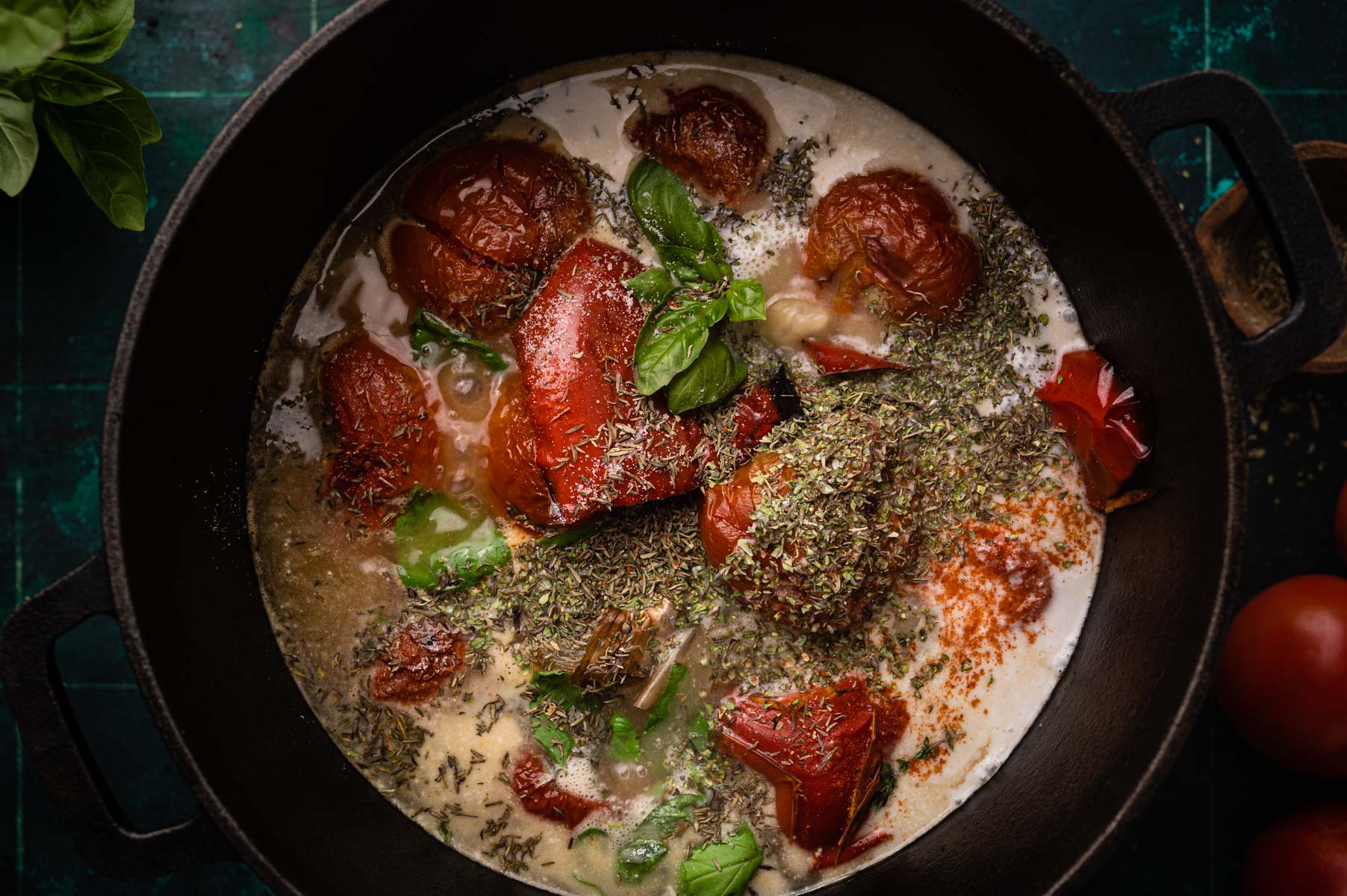A skillet with roasted tomatoes and herbs in a creamy sauce, surrounded by fresh ingredients on a dark surface.