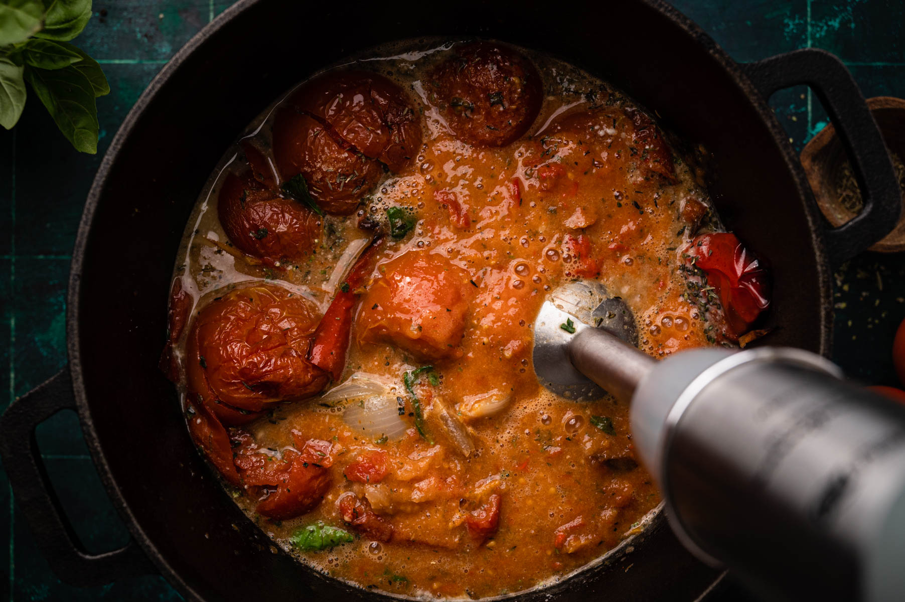 A skillet with simmering tomato sauce and roasted tomatoes, garnished with herbs, being stirred with a spoon.