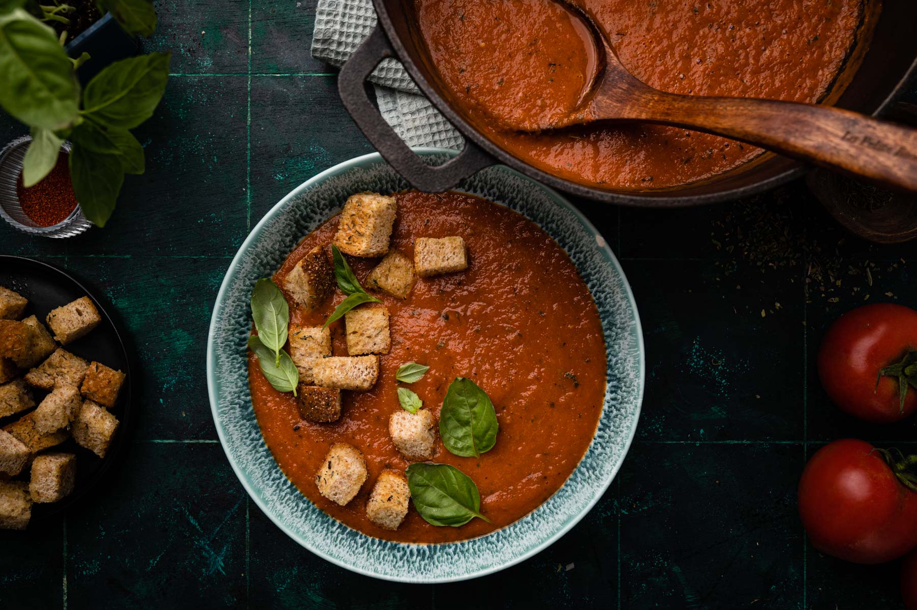 A bowl of tomato soup with croutons and basil leaves, accompanied by a wooden spoon and fresh tomatoes on a dark surface.
