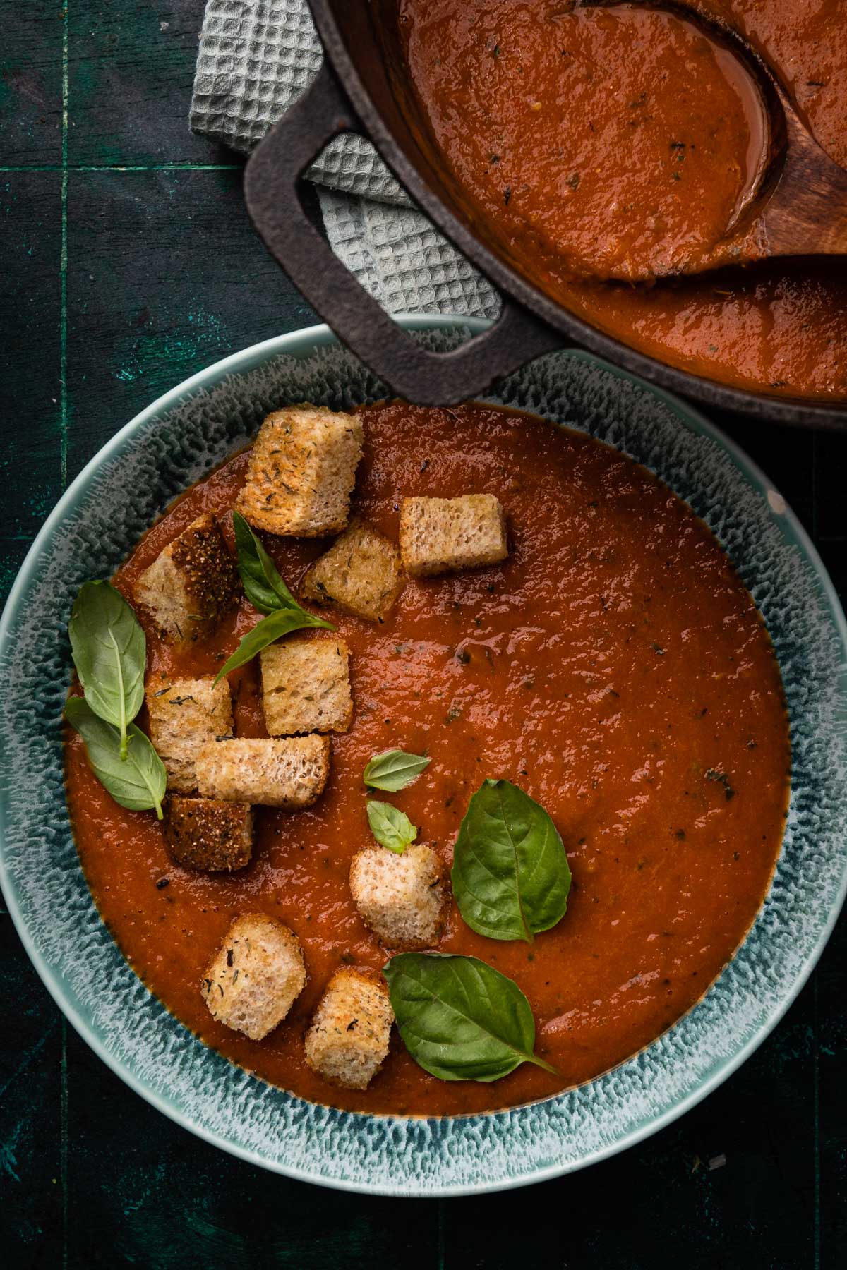 A bowl of tomato soup garnished with basil and croutons, next to a cast iron pot of soup and a cloth, on a dark wooden table.