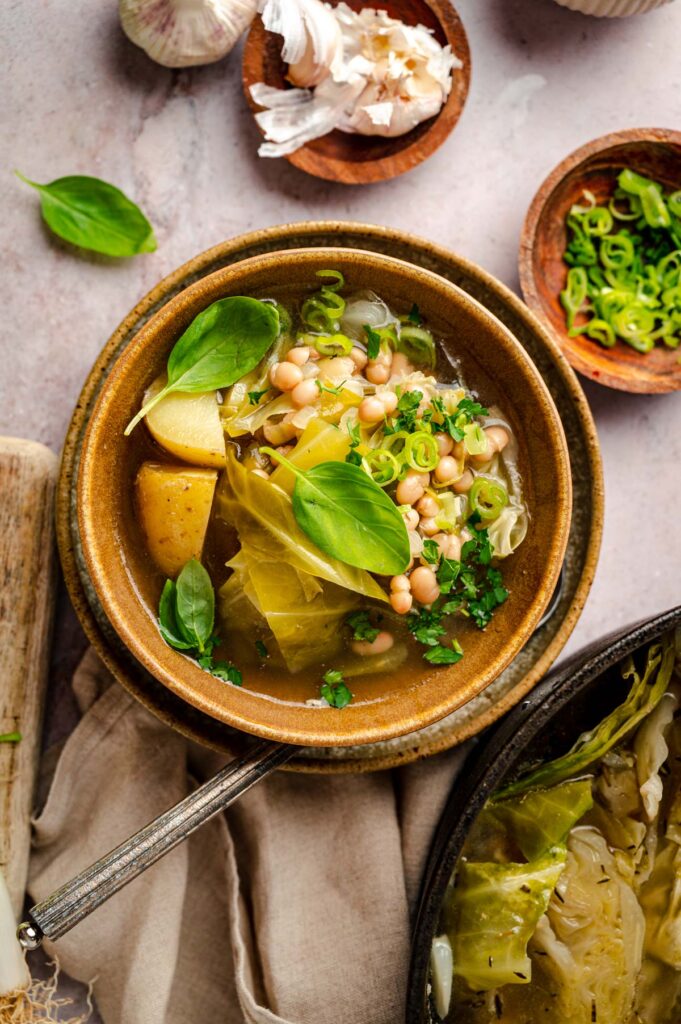 A bowl of vegetable soup with potatoes, beans, cabbage, green onions, and basil. Nearby are garlic bulbs, peeled garlic, a small bowl of green onions, and a pot with cooked cabbage.