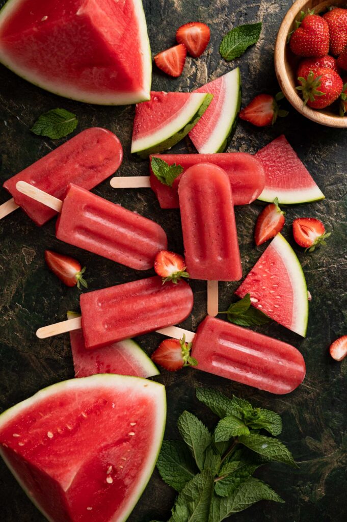 Assorted watermelon and strawberry popsicles are displayed on a dark surface surrounded by fresh watermelon slices, halved strawberries, and mint leaves.
