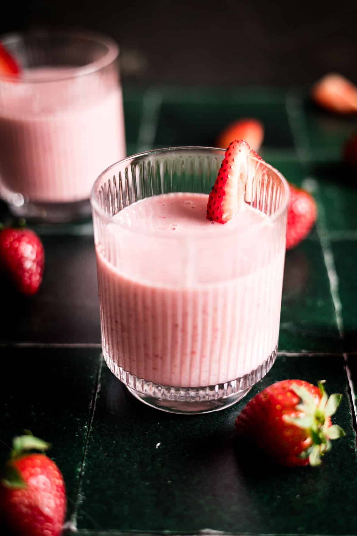 Pink strawberry with banana smoothie in a glass cup on green tile counter with fresh strawberries.