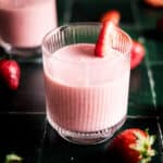 Pink strawberry with banana smoothie in a glass cup on green tile counter with fresh strawberries.