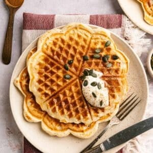 Several protein waffles on a plate with vegan whipped cream and pumpkin seeds.